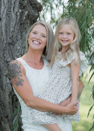 A smiling blonde woman with arm tattoos holds a young girl near a tree outdoors.