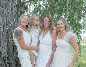 Four women in white dresses smile together outdoors near a large tree, including a young girl being held.