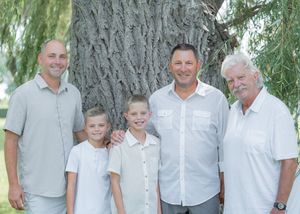 Four generations of men and boys pose smiling outdoors near a large tree on a sunny day.
