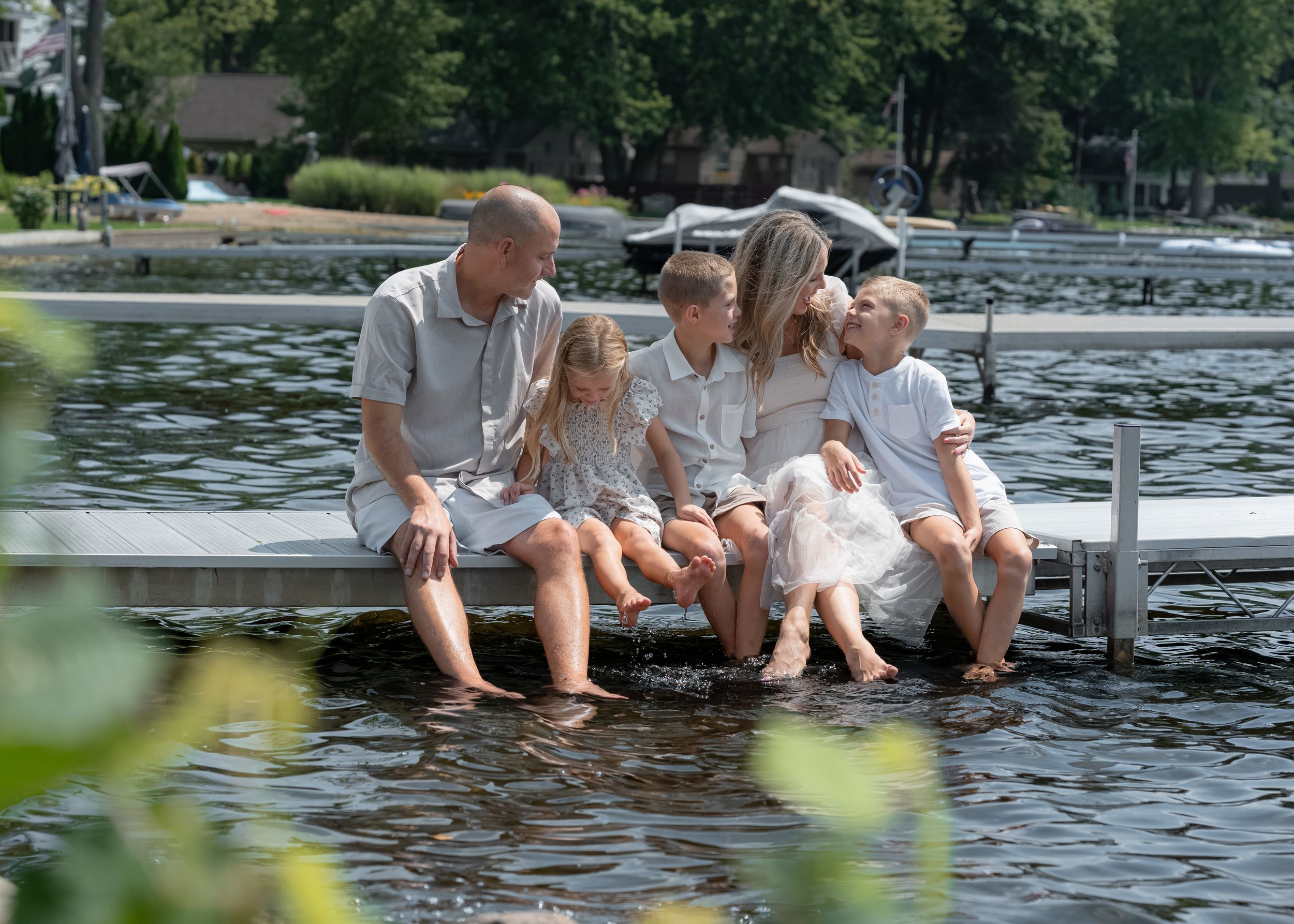 Family of five sitting on a dock with feet in water, surrounded by lush trees and a serene lake backdrop.