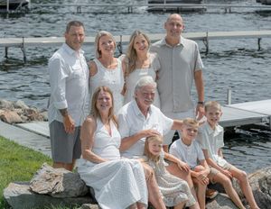 Multi-generational family posing together on rocky lakeside dock in coordinated white and grey outfits on a sunny day.
