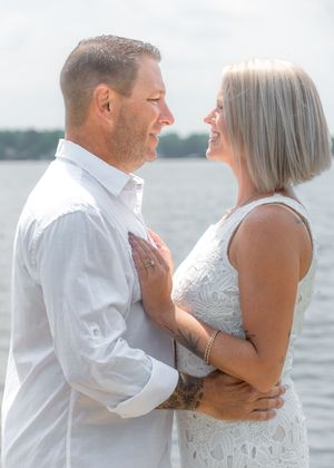 Couple embracing and smiling at each other near a waterfront, woman in white lace dress, man in white shirt.