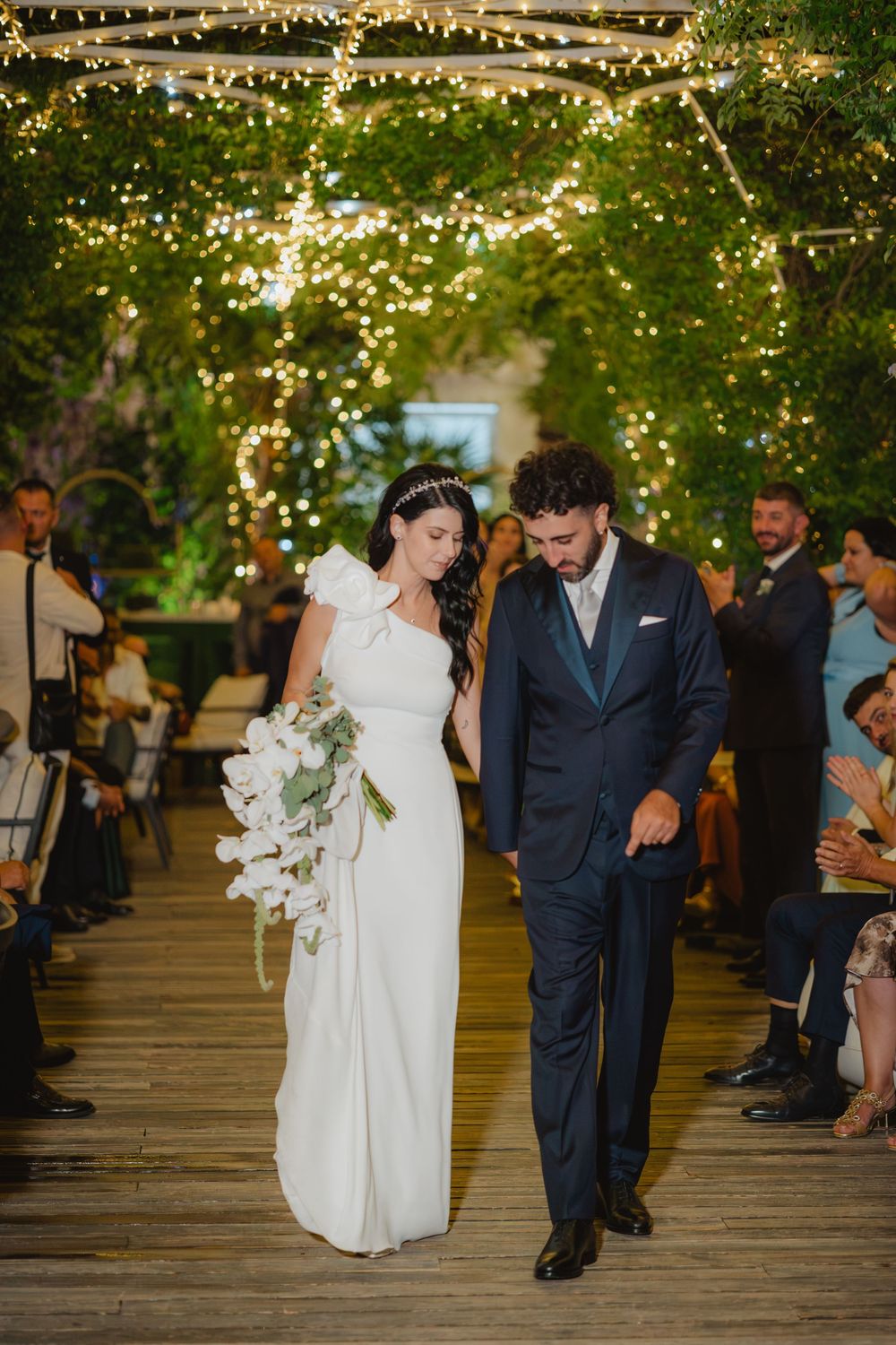 Lorenzo e Laura camminano insieme sotto le luci e la vegetazione del ricevimento.