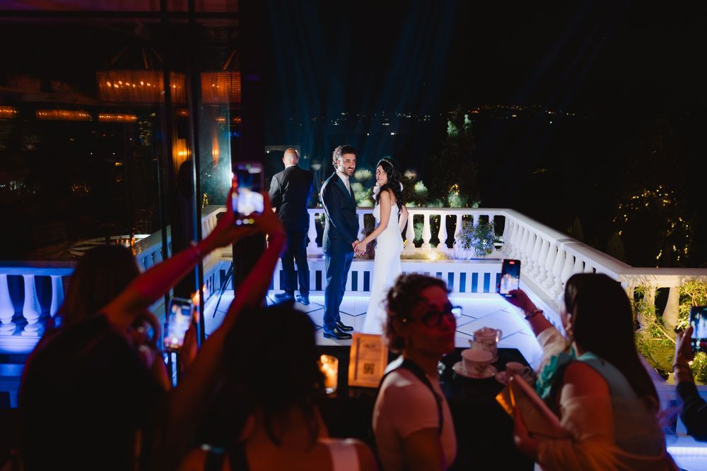 Gli sposi festeggiano all’aperto di sera, davanti agli invitati e ai telefoni alzati.