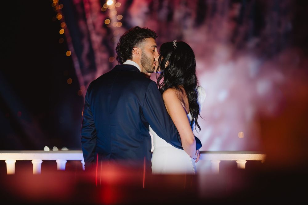 Lorenzo e Laura si avvicinano sotto le luci della sera, con riflessi in primo piano.
