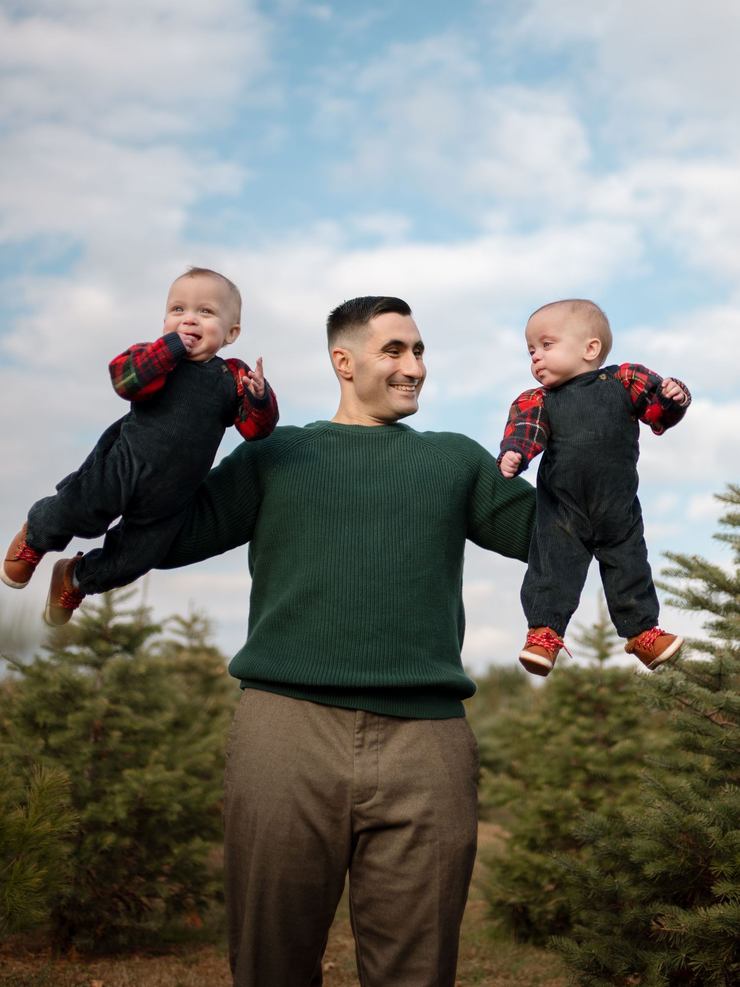 Smiling father in a green sweater lifts two giggling twin babies in plaid and overalls at a Christmas tree farm under blue sky.