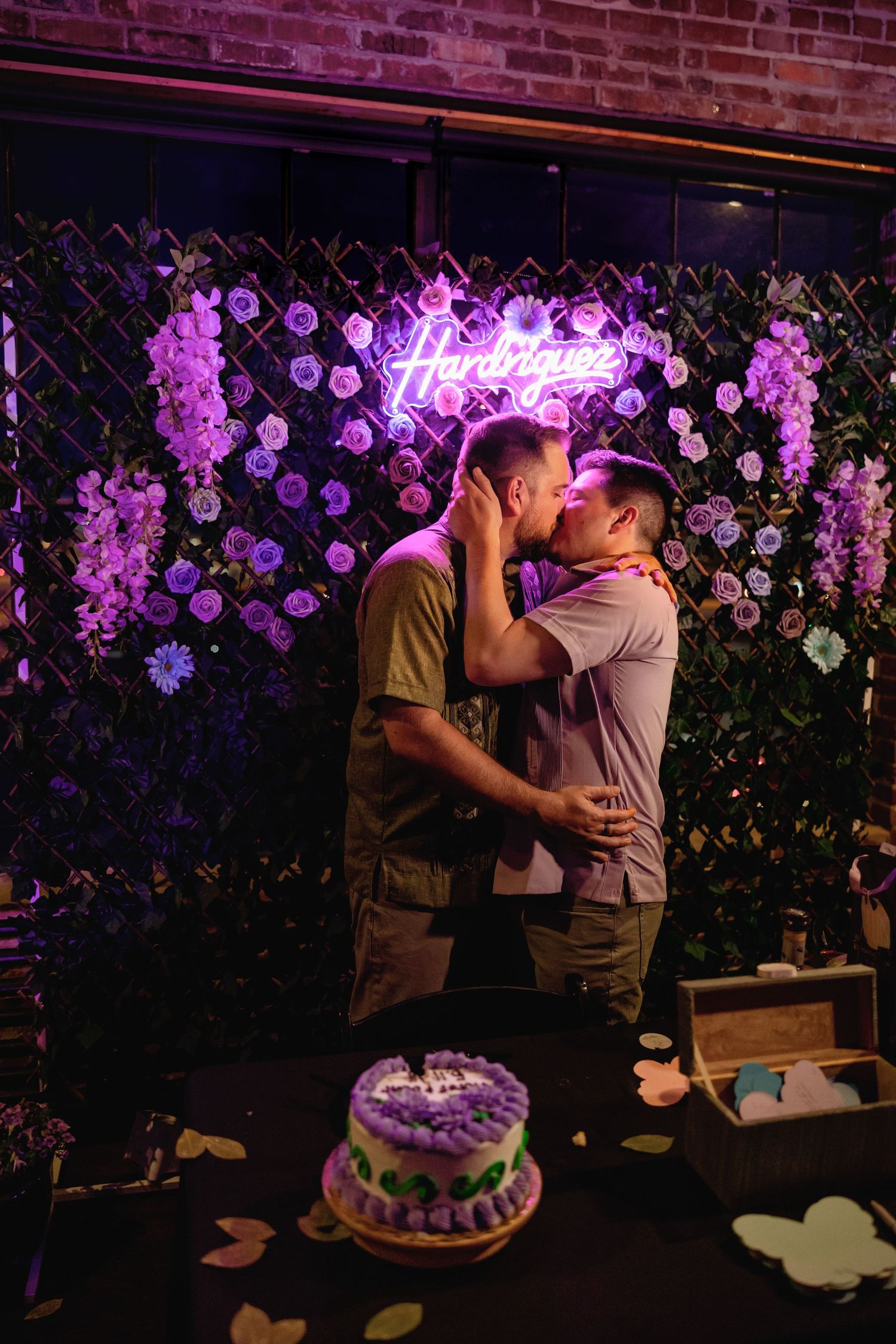 Two men share a romantic kiss in front of a purple neon sign and floral backdrop with a decorated cake nearby.
