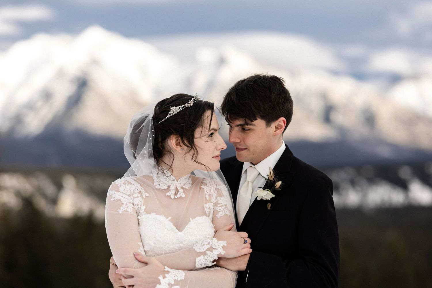 Bride and groom embrace with snowy mountain peaks behind them in a romantic winter wedding portrait.
