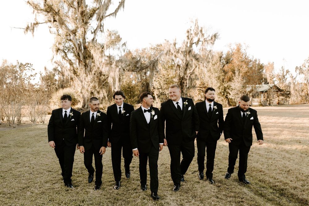 Groom and groomsmen in black tuxedos walking outdoors at a wedding venue with Spanish moss trees.