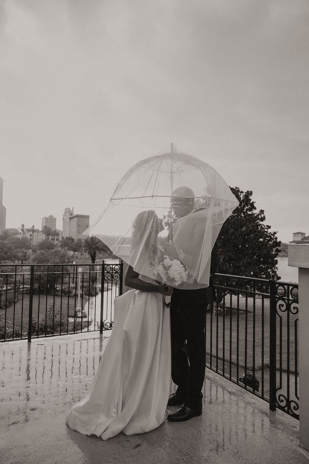 Black and white photo of a bride and groom sharing an umbrella on a rainy wedding day on an outdoor balcony.