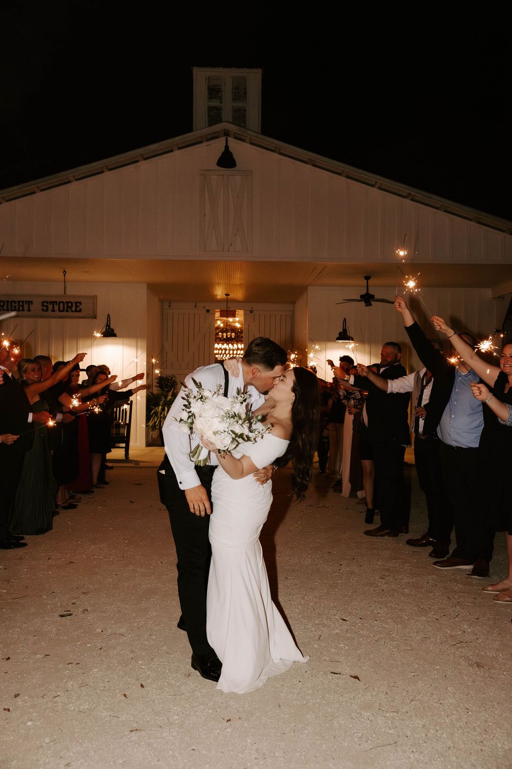 Bride and groom share a romantic dip kiss during sparkler exit at night outside a white barn wedding venue.
