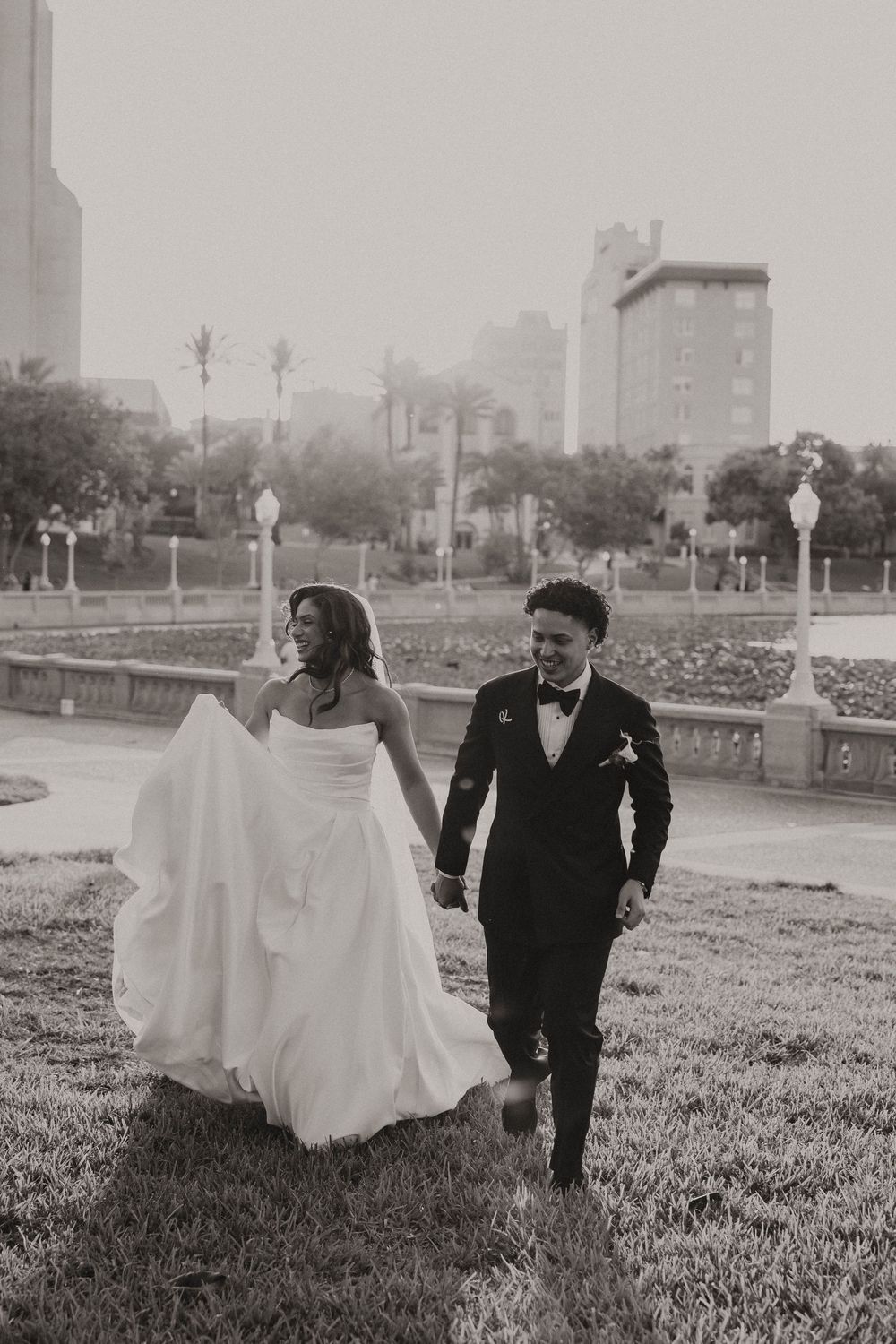 Black and white photo of a bride and groom holding hands, walking through a park with city buildings in the background.