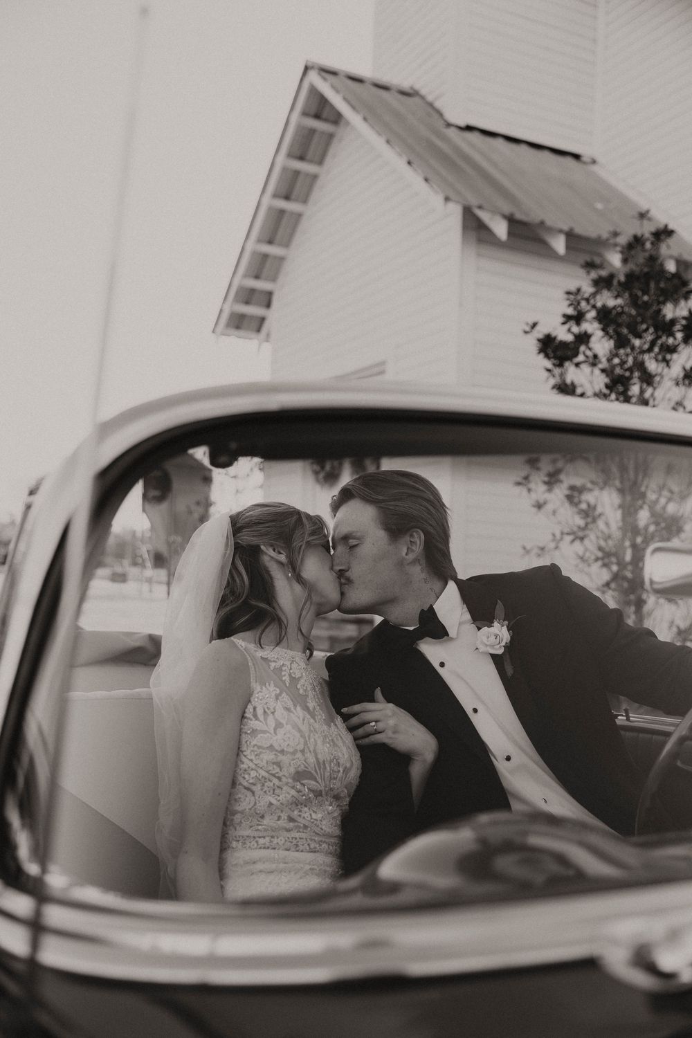 Black and white photo of a bride and groom sharing a kiss inside a vintage car outside a white church.