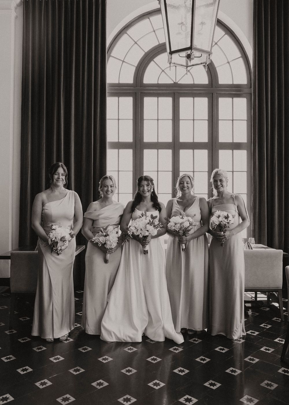 Black and white photo of a bride and four bridesmaids in elegant gowns holding bouquets before a grand arched window.