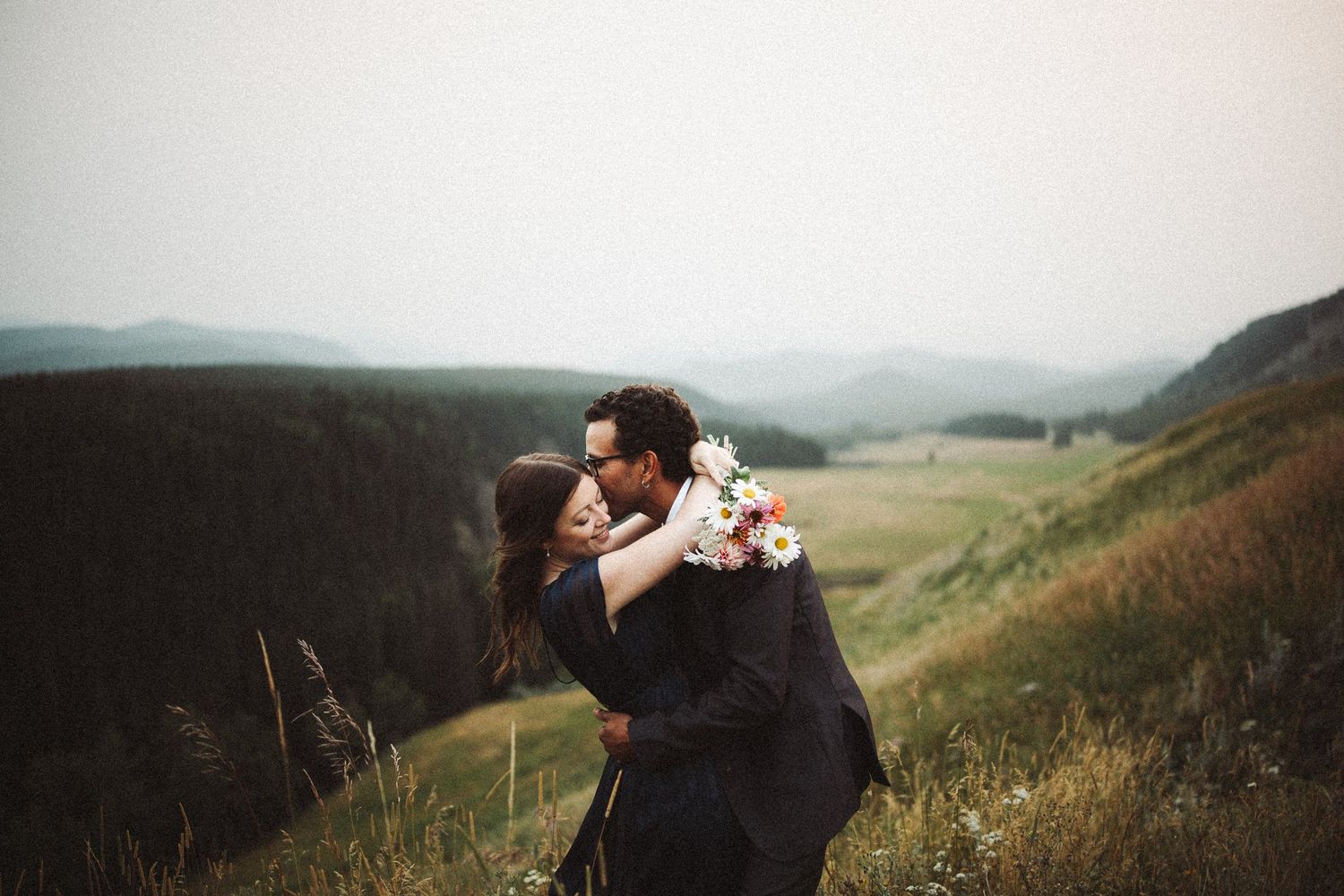 Engaged couple embracing with a wildflower bouquet on a grassy hillside above a mountain valley