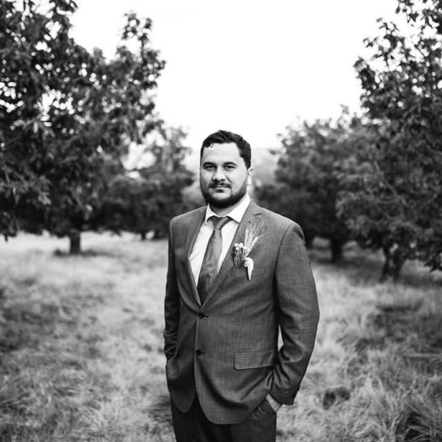 A groom in a gray suit with boutonniere stands in an orchard, black and white wedding portrait photo.
