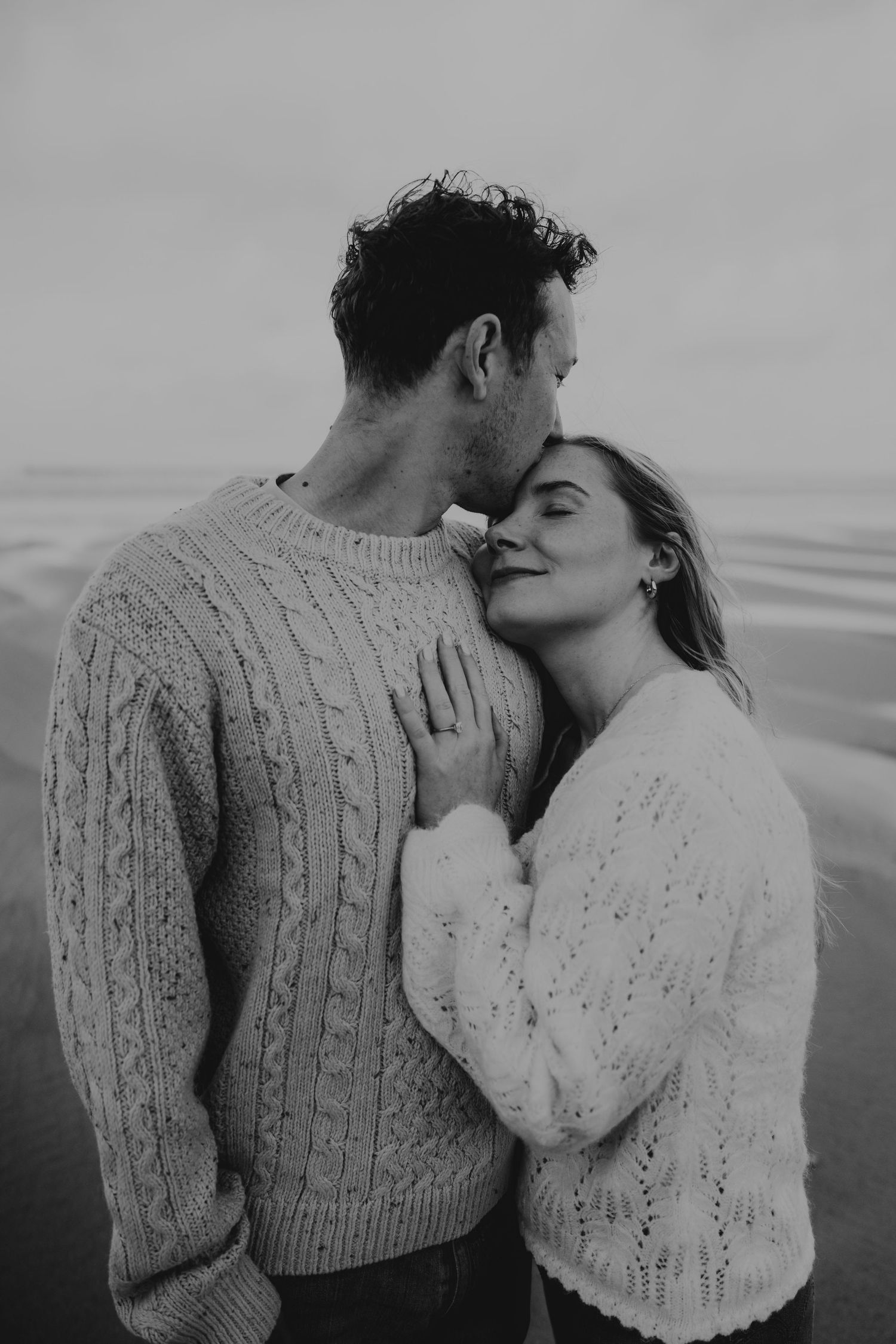 Black and white photo of a romantic couple embracing at watergate bay, both wearing cozy knit sweaters.