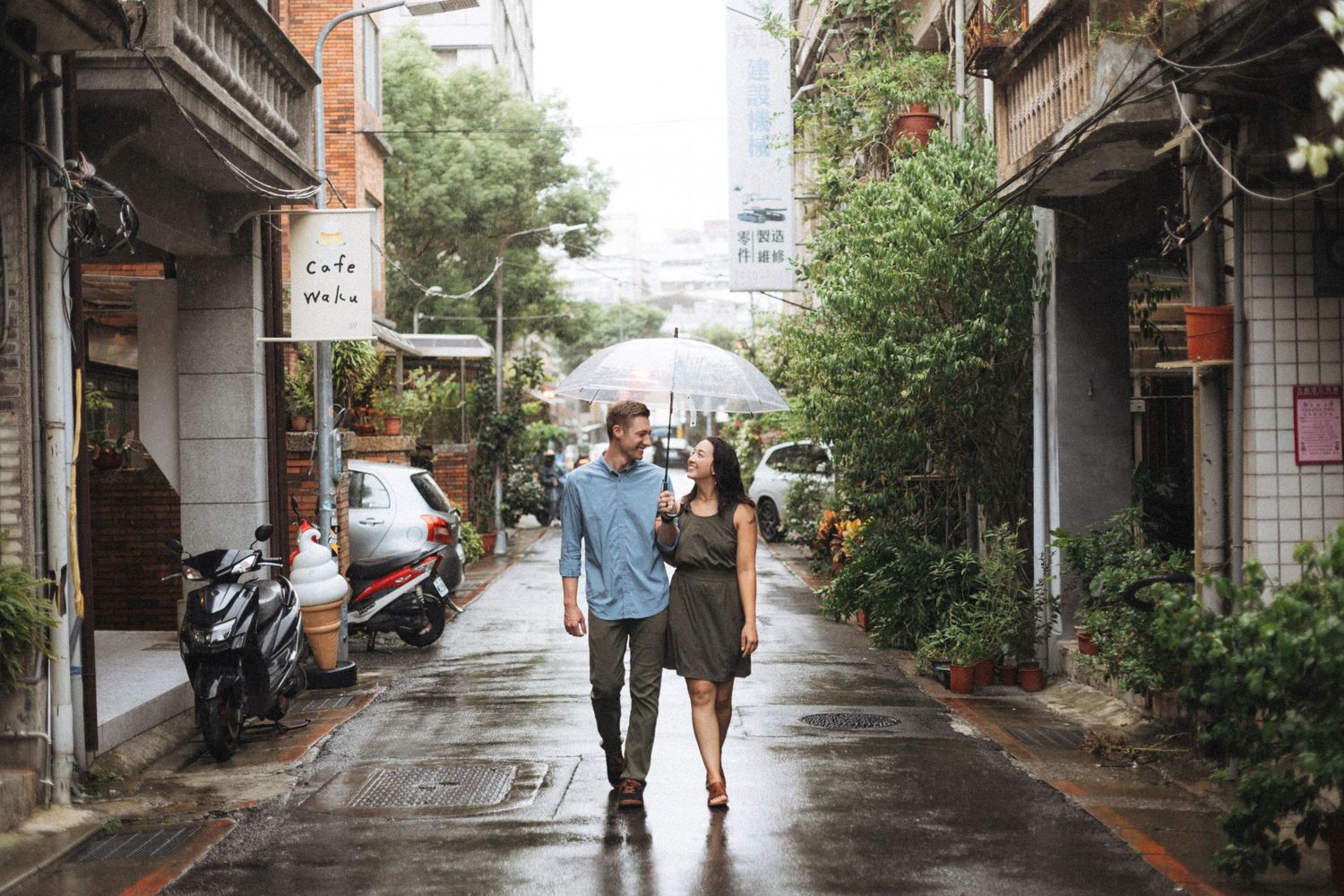 Couple walking hand in hand on a rainy urban alley lined with shops and greenery.
