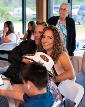 A couple smiles and poses together at an indoor event, with the woman holding a white cowboy hat, surrounded by guests.