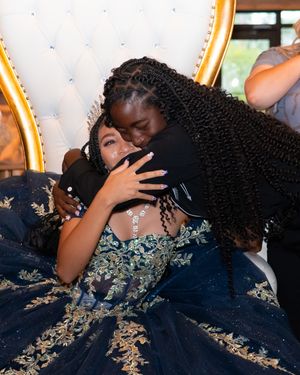 Two girls share a warm hug at a quinceañera, one wearing an elegant navy blue and gold embroidered ball gown.