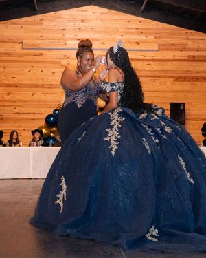Two women in navy blue quinceañera gowns with gold lace share a tender moment at an elegant celebration venue.