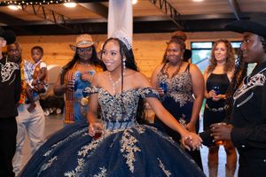 Young woman in navy blue quinceañera ball gown with gold embroidery and crown celebrates with guests at elegant party.