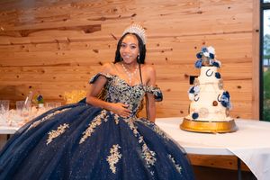 Young woman in navy blue and gold quinceañera gown wearing a tiara poses beside a decorated white cake.