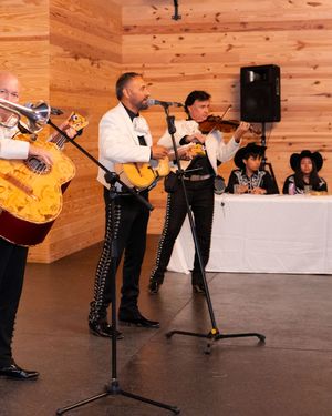 Mariachi band performs with guitar, trumpet, and violin at an indoor event venue with wood panel walls.