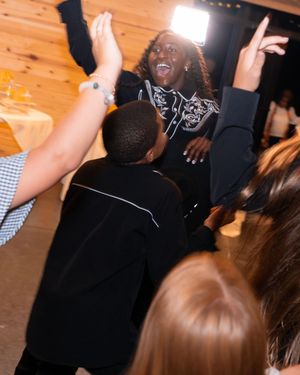 Joyful woman in black outfit celebrates with raised hands surrounded by guests at a lively indoor party.