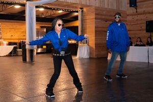 Two people in matching blue jackets dance on an indoor wooden floor at an event venue.