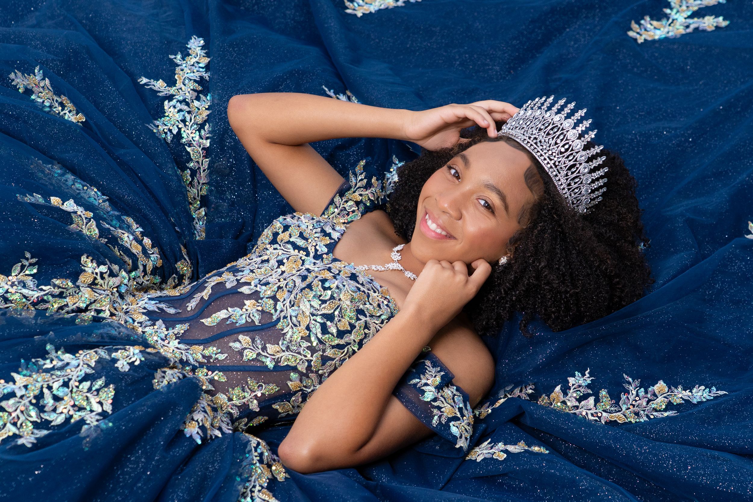 Young woman in navy blue embroidered gown and crystal tiara poses elegantly for a quinceañera or pageant portrait.