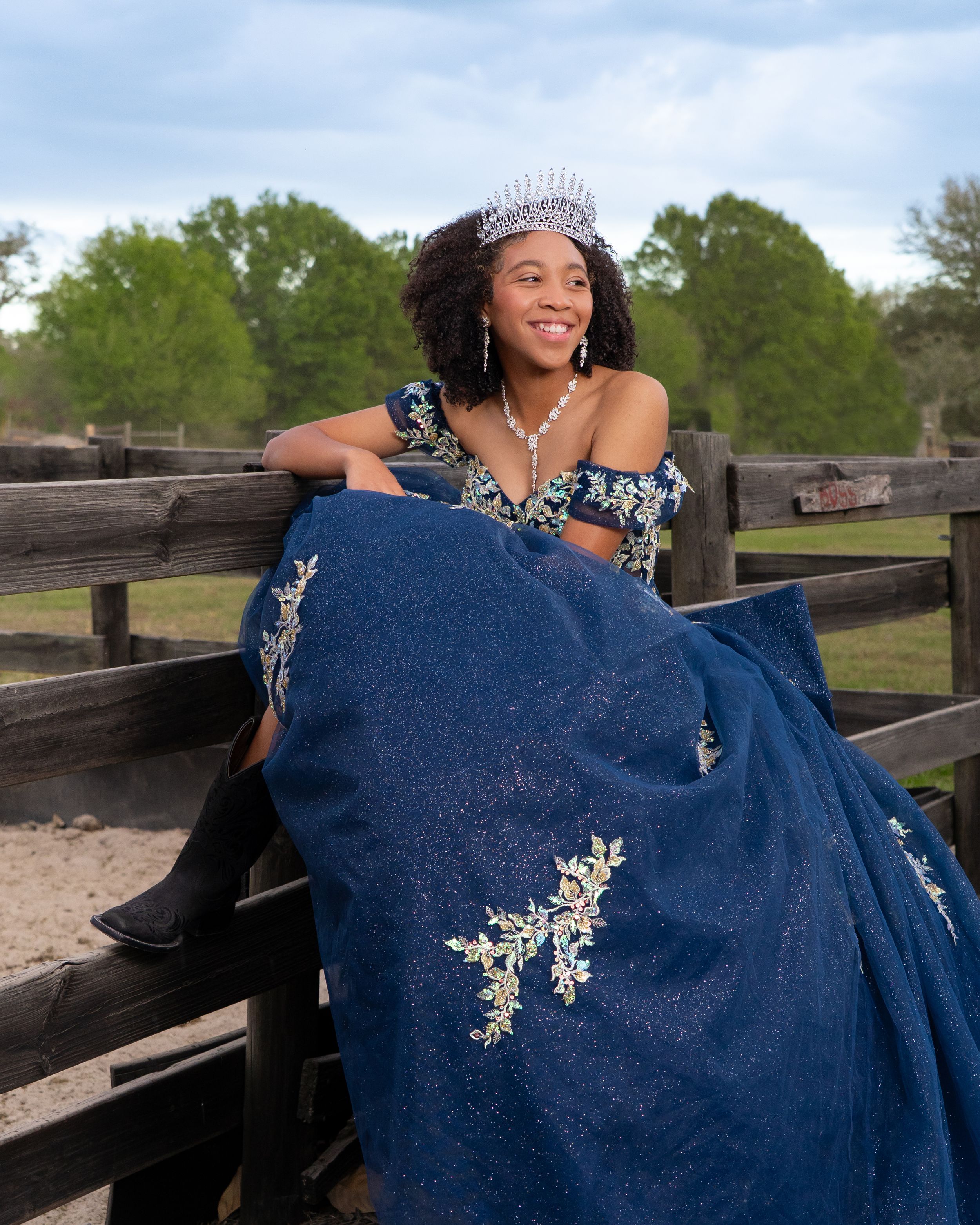 Young woman in navy blue ball gown with silver embroidery and tiara poses on rustic wooden fence outdoors.