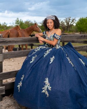 A young woman in a navy blue quinceañera ball gown with silver embroidery and tiara poses by a wooden fence with horses.