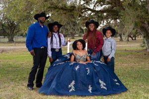 Family in cowboy hats posing outdoors with girl in blue quinceañera gown in a park setting.
