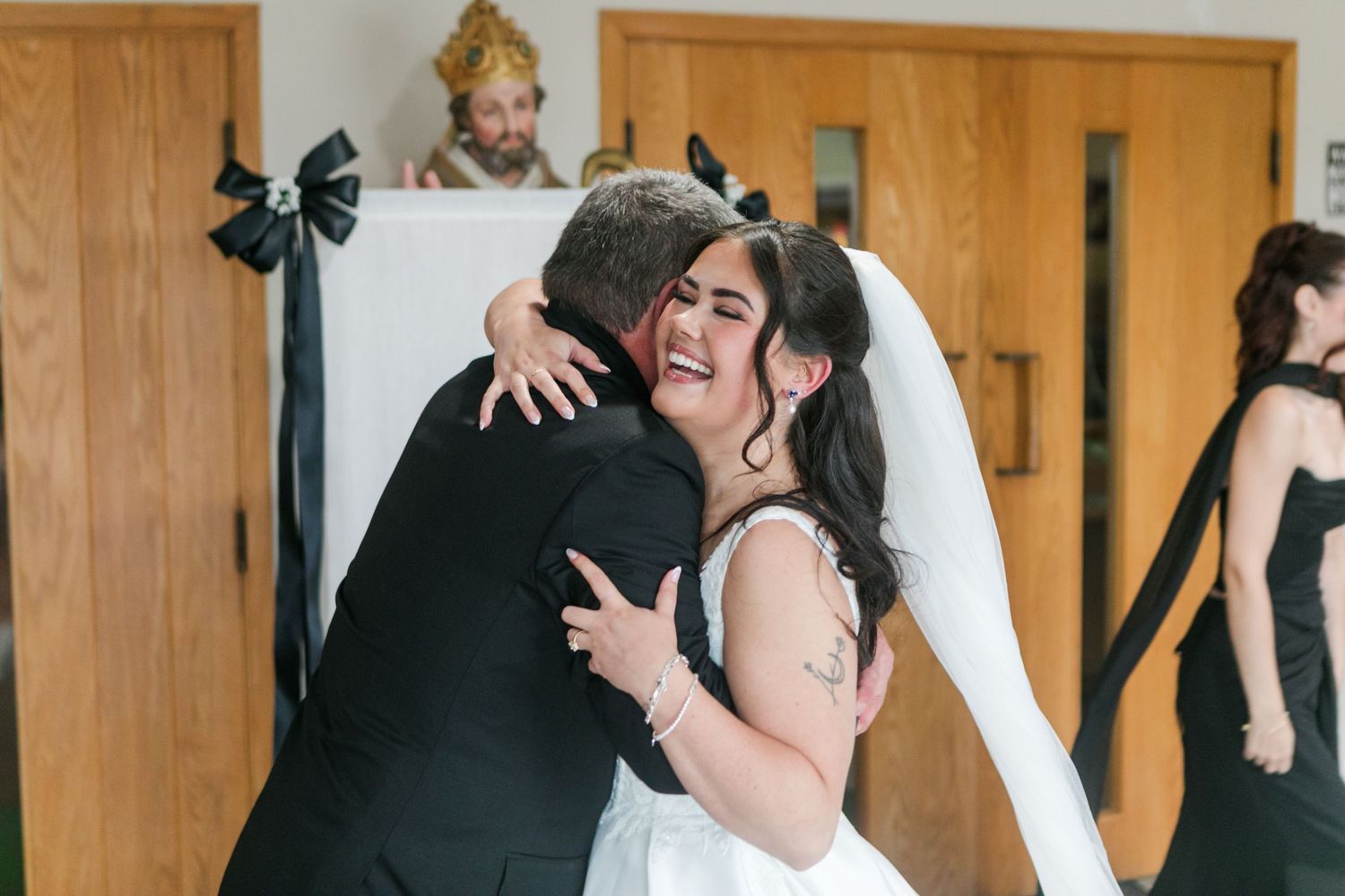 Smiling bride in a white wedding dress and veil hugs a man in a dark suit inside a church with wooden doors.