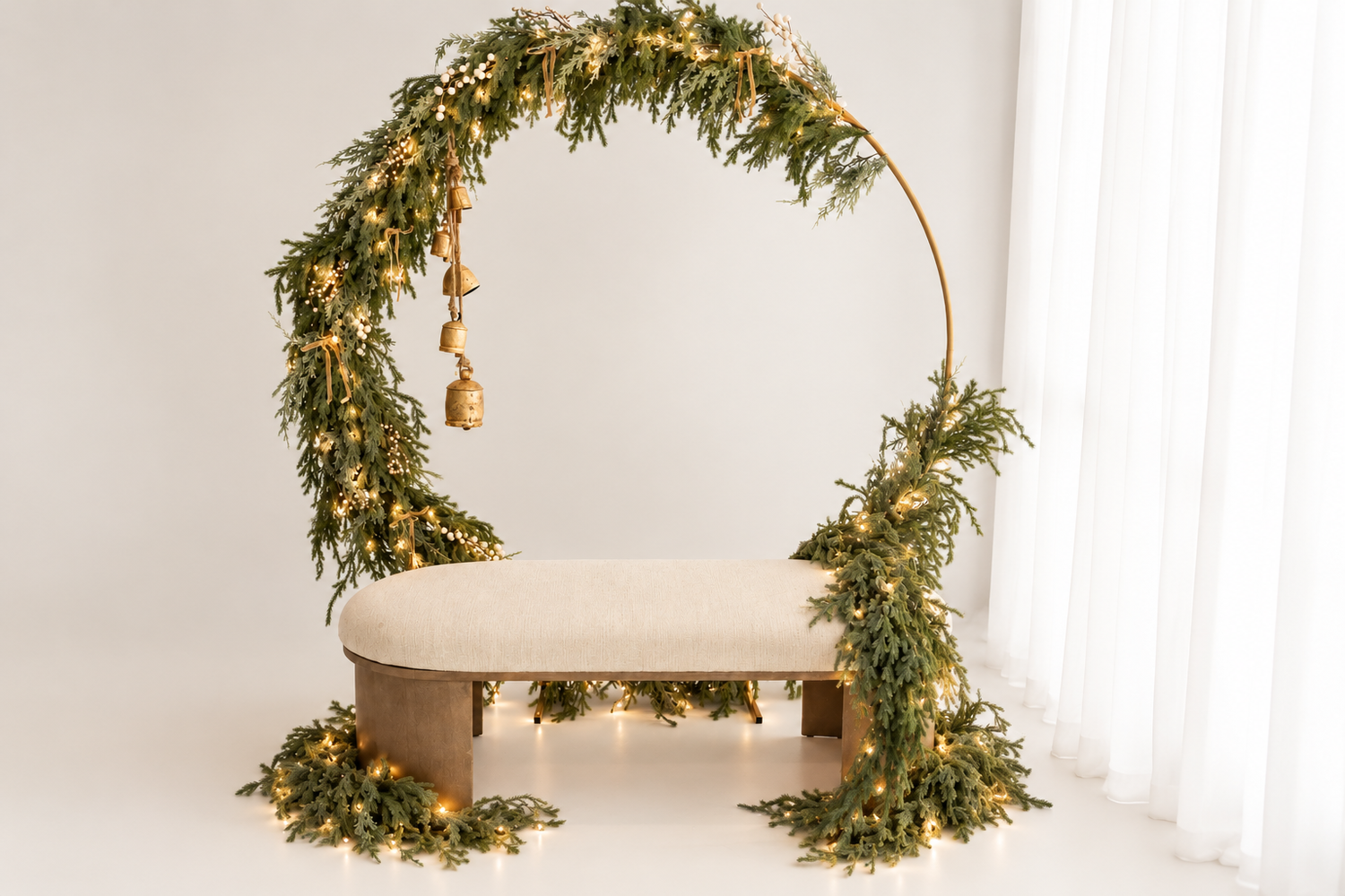 Circular greenery arch with gold bells and fairy lights surrounding a cream upholstered bench, a festive holiday photo studio backdrop.