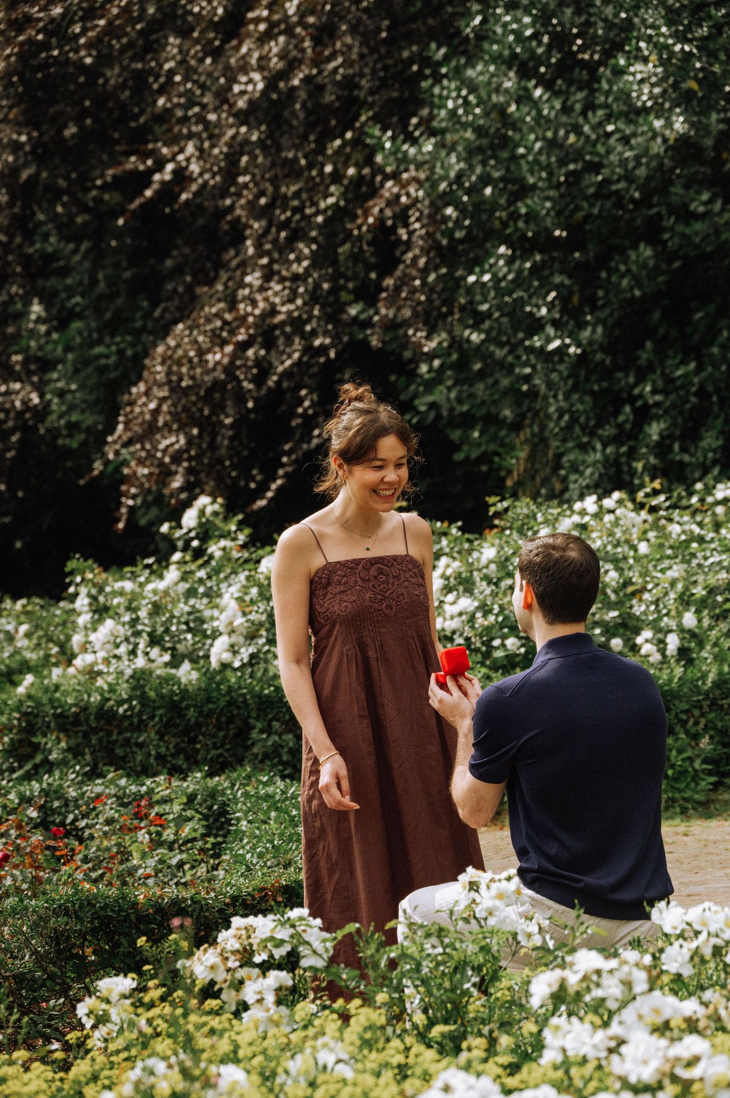 Man in a navy shirt kneels holding a red ring box as a smiling woman in a brown dress reacts during a marriage proposal in Vondelpark.