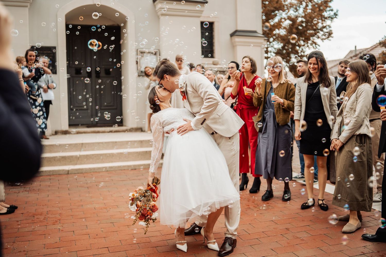 Bride and groom share a kiss on church steps as joyful wedding guests surround them and bubbles float through the air.