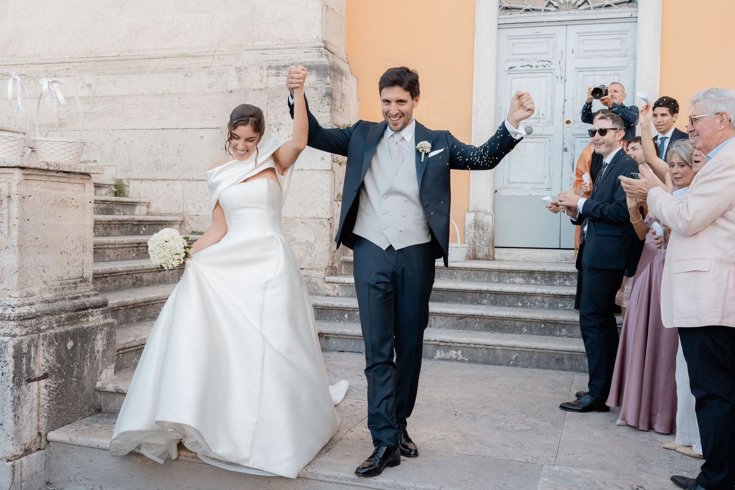 A joyful bride in a white gown and groom in a dark suit raise their hands as wedding guests cheer on stone steps outside a church.