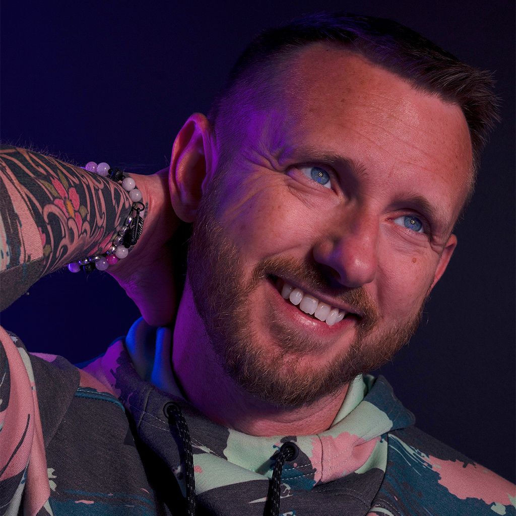 Smiling bearded man with blue eyes and a tattooed arm raised behind his head, wearing a camouflage-print hoodie against a purple backdrop.