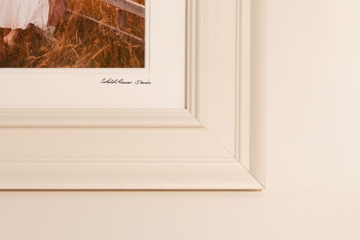 Bottom corner of a cream white wooden picture frame showing a signed fine art photograph of golden autumn grass and a figure in white.