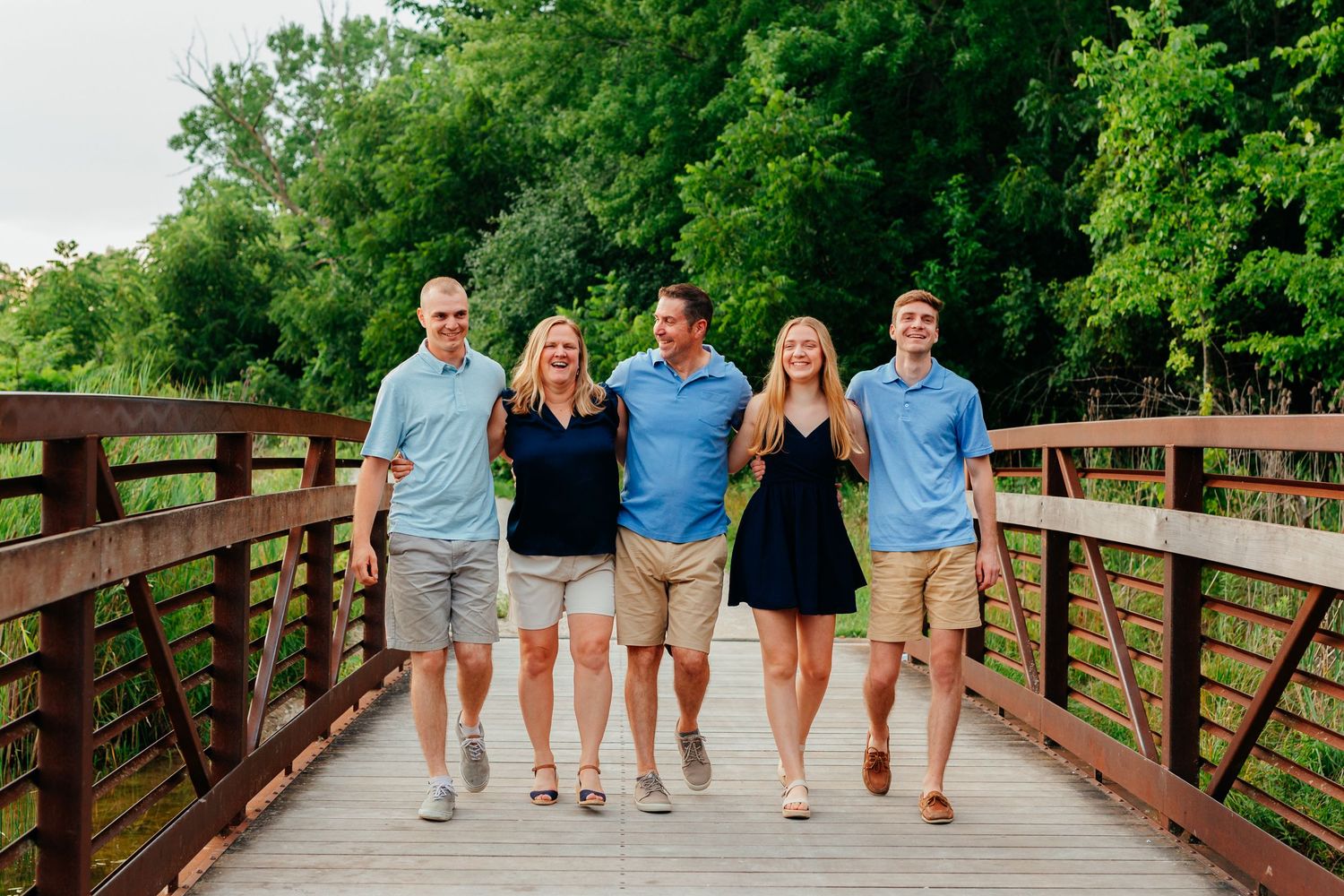 Family of five in blue and black outfits walks arm in arm across a wooden bridge surrounded by lush green trees.