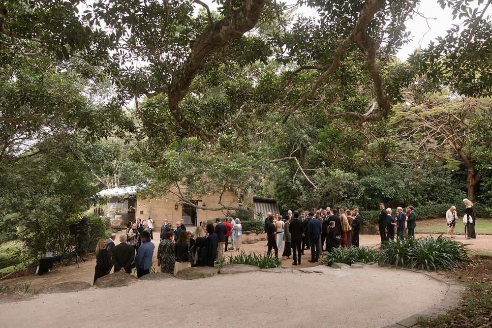 Wedding guests in formal attire gather outdoors beneath large shady trees near a stone building along a curving gravel path.