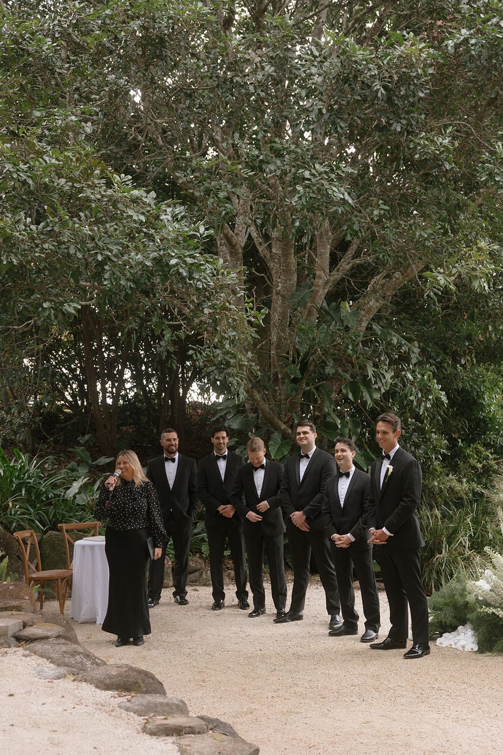 Groomsmen in black tuxedos stand along a gravel path beneath large trees while an officiant in black speaks at an outdoor wedding ceremony.