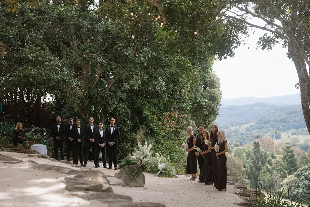 Groomsmen in black suits and bridesmaids in dark dresses line a garden path at a hilltop wedding ceremony overlooking green valley views.