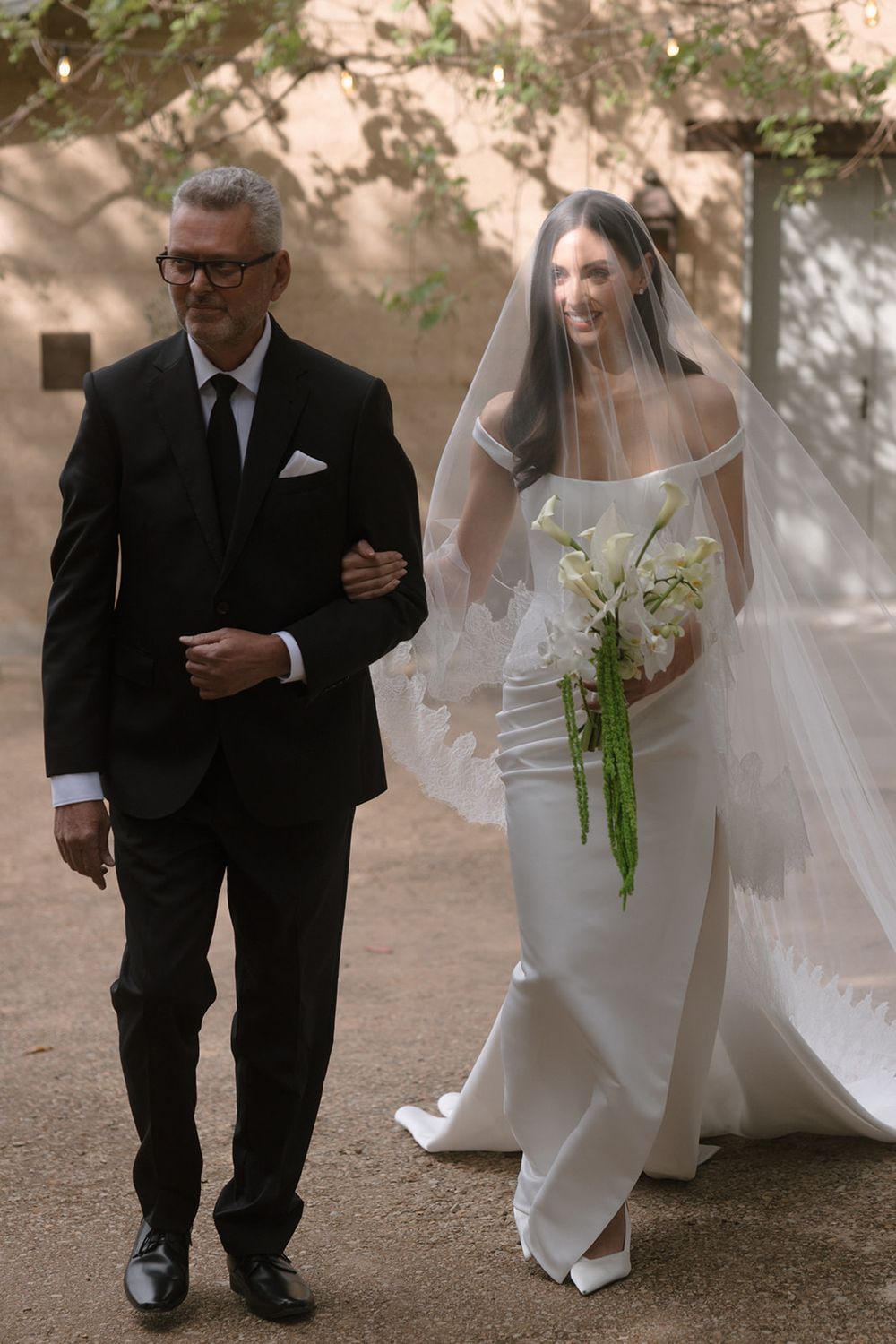 A bride in a fitted white gown and long veil holds a calla lily bouquet as a man in a black suit walks her down a sunlit path.