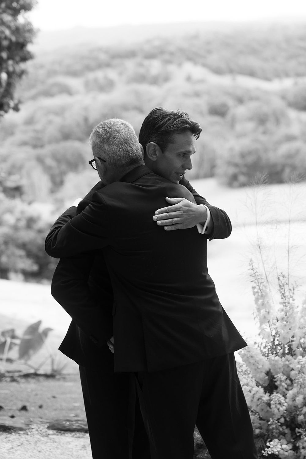 Two men in dark suits share a heartfelt embrace outdoors in a black and white photo, with a soft hillside and flowers behind them.