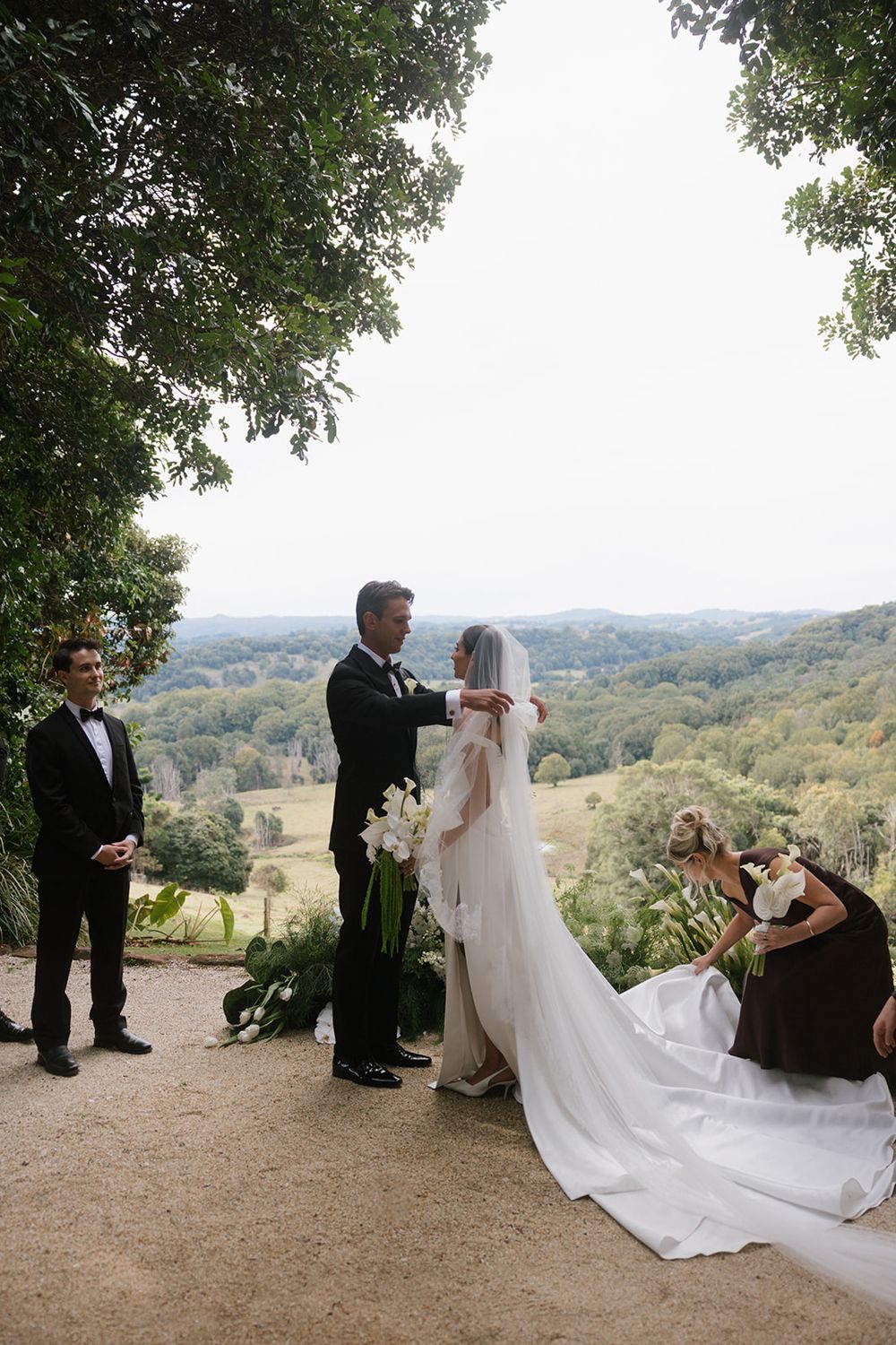 A groom in a black tuxedo adjusts his bride's long veil and white gown on a hilltop with sweeping countryside views as bridesmaids assist.