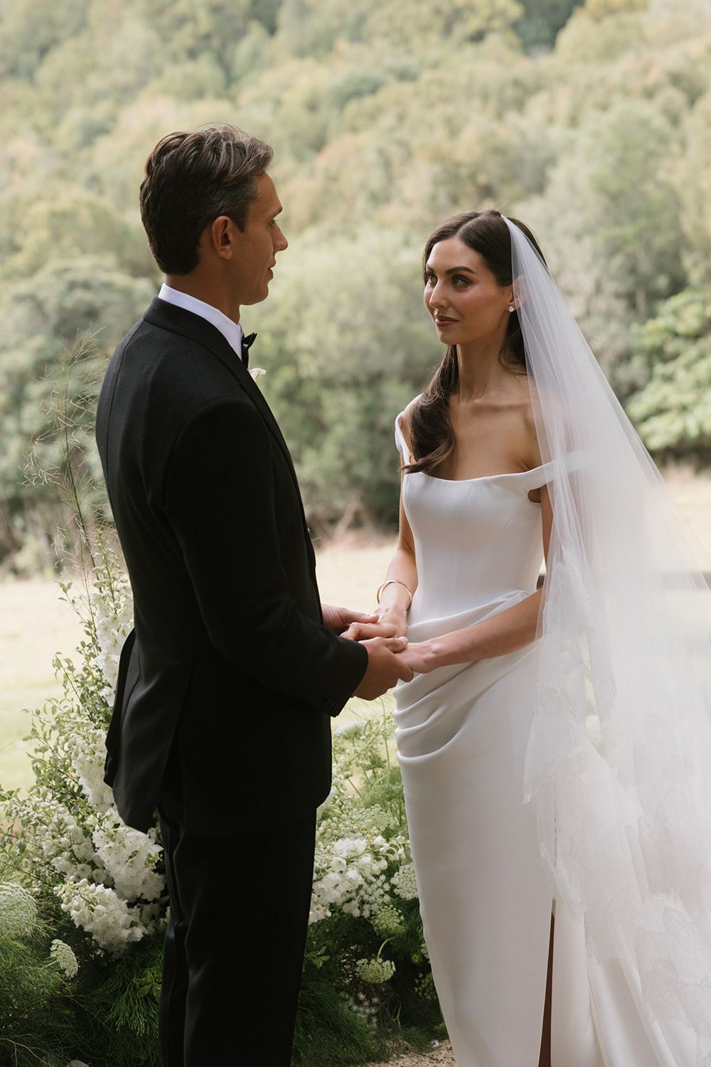 Bride in an off-shoulder white gown and long veil holds hands with a groom in a black tuxedo during an outdoor garden wedding ceremony.