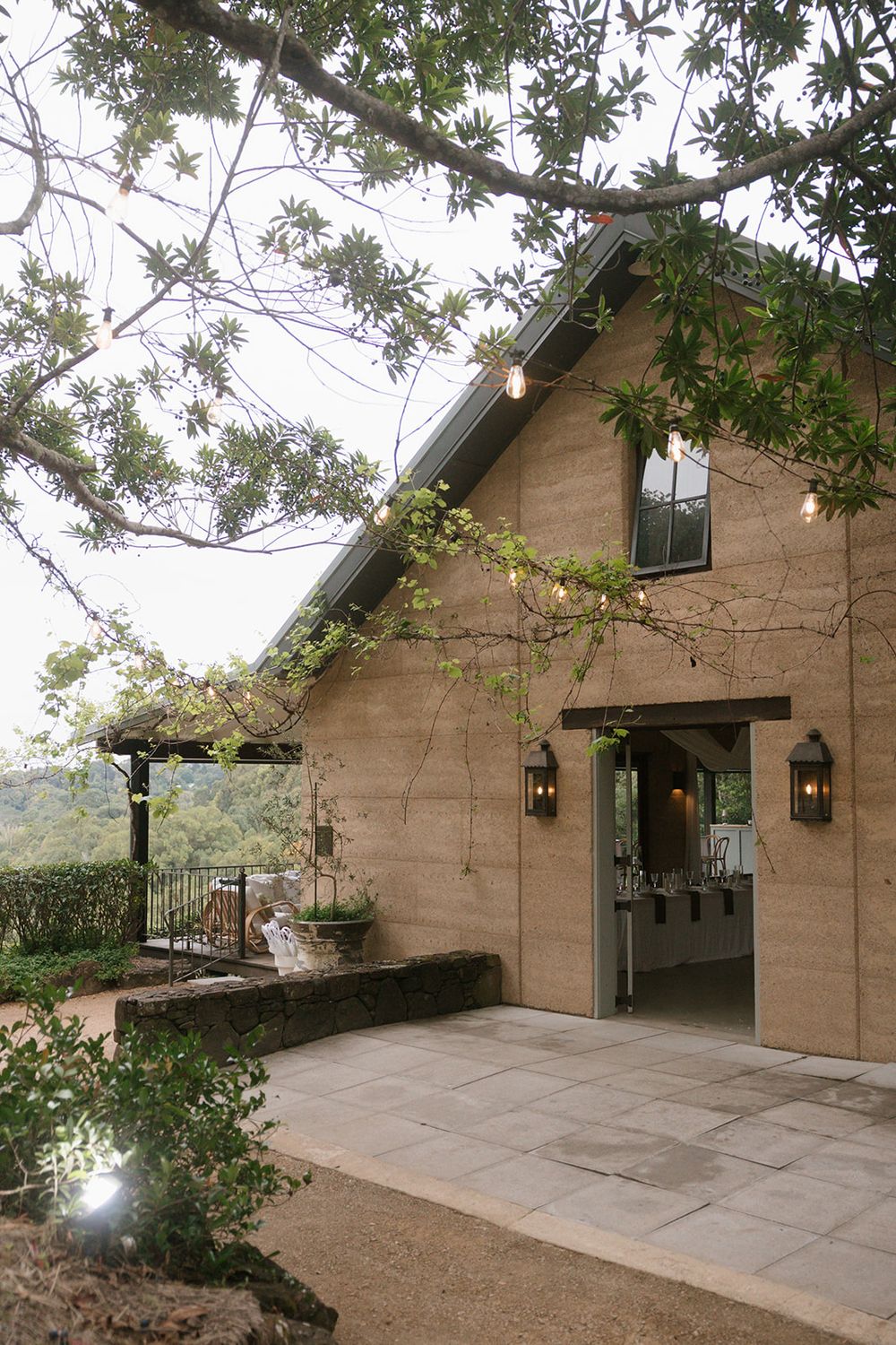Rustic stone barn venue with open doorway, glowing lantern sconces, and a paved patio beneath tree branches draped with string lights.